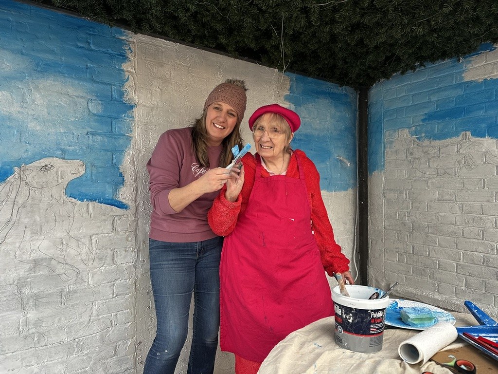 Margaret, the Pink Patsy illustrator, with paintbrushes in hand in front of the first pencil sketch and blue sky of the Crocodile Café mural