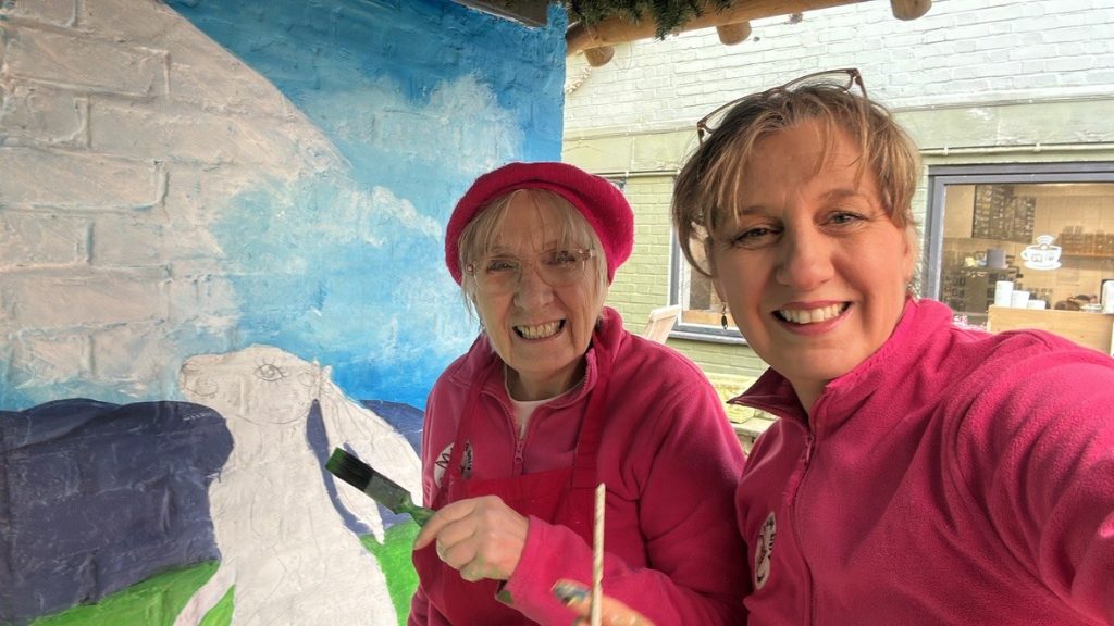 Margaret and a member of the Pink Patsy team smiling beside the mural as the sky and hills are painted in