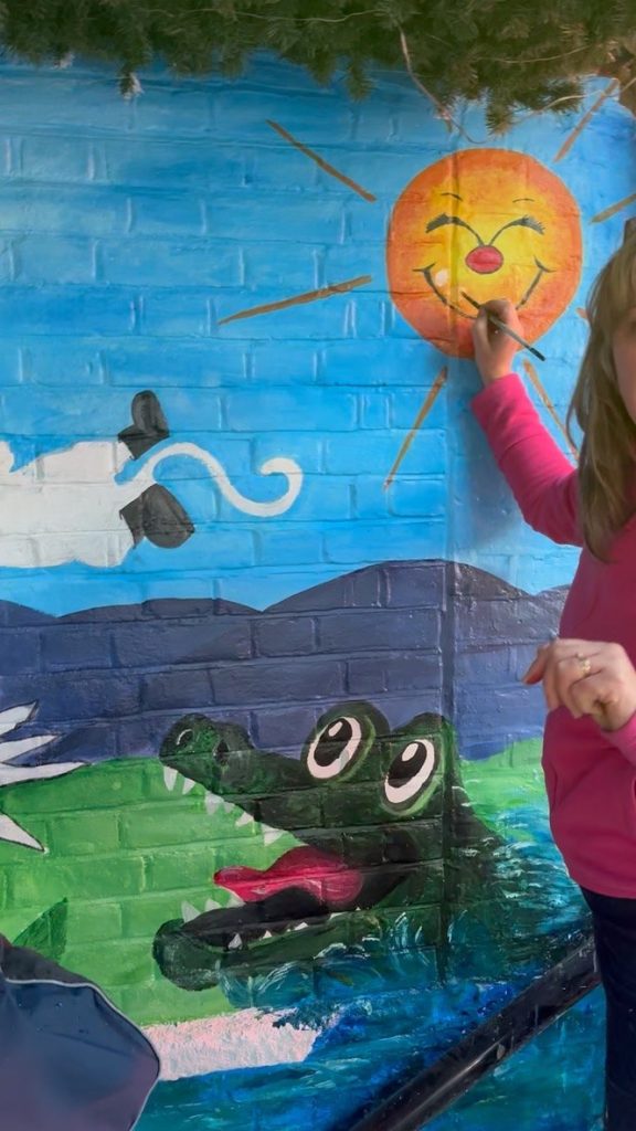 Painting the smiling sun above a grinning green crocodile on the Pink Patsy mural at the Crocodile Café