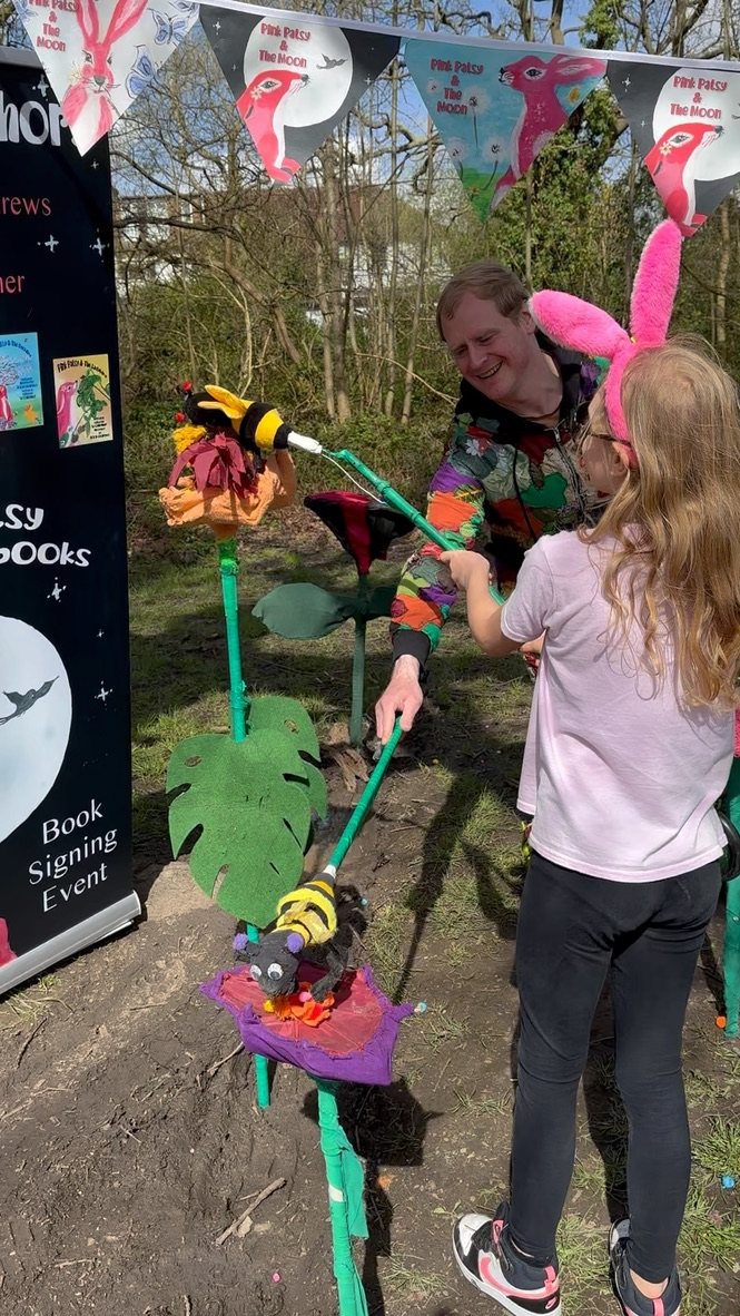 Jake in his colourful costume helping a girl in pink bunny ears use a bee puppet to carry pollen between giant flowers at a Pink Patsy event