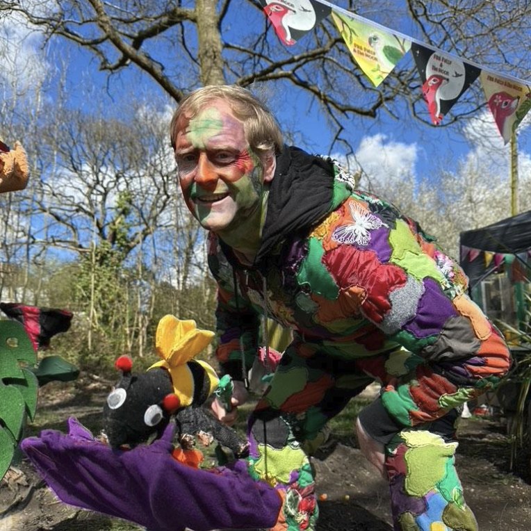 Jake in a colourful patchwork costume with his bee puppet on a purple flower, showing how bees pollinate at Pink Patsy World Bee Day in Swan Lane Open Space, Whetstone