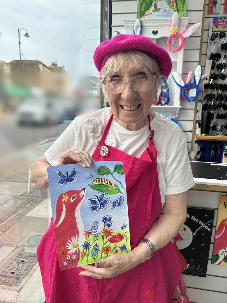 Margaret, the Pink Patsy illustrator, smiling in her pink beret and apron outside the Magmay Books shop on Muswell Hill Broadway, holding her finished painting of butterflies, a caterpillar and wildflowers