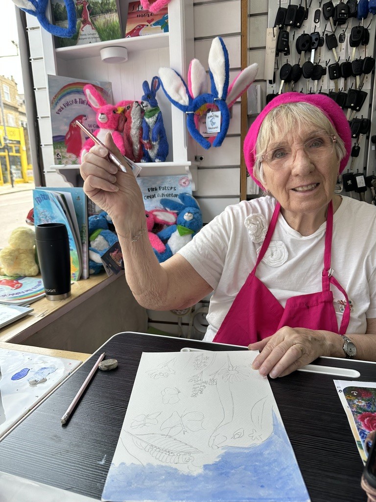 Margaret, the Pink Patsy illustrator, starting a new painting in the front window of the Magmay Books shop in Muswell Hill, with the pencil sketch and first wash of blue sky