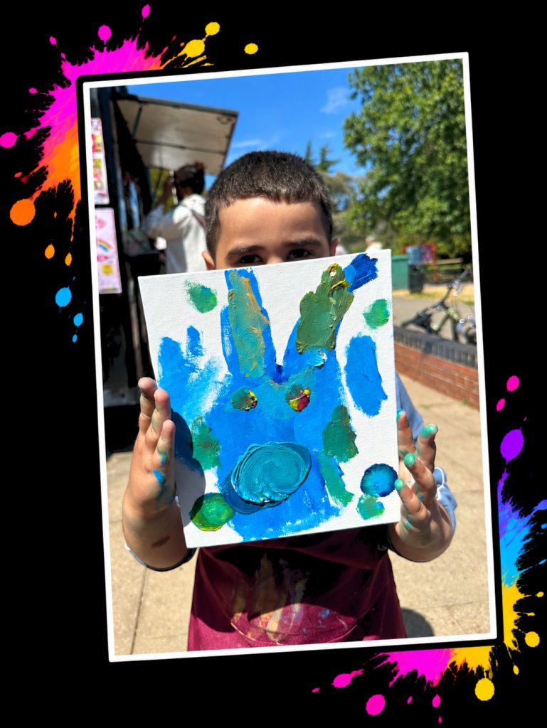 A young artist with painty fingers holding up his blue Bobby Blue painting at a Painting in the Park event