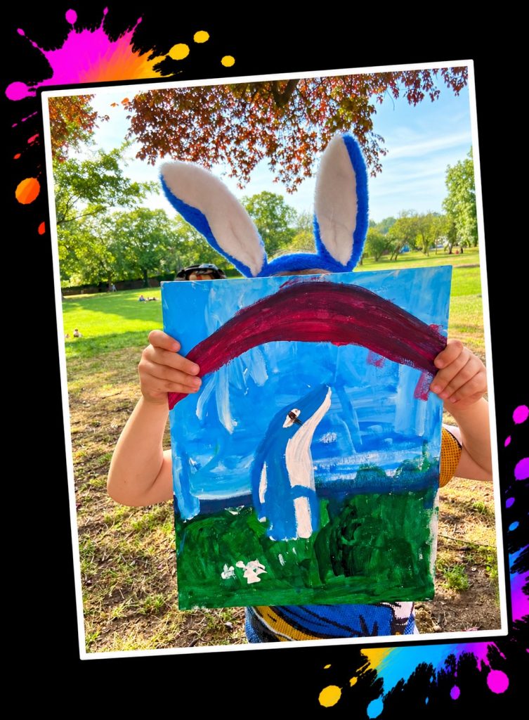 A young artist in Bobby Blue bunny ears holding up a painting of Bobby Blue under a rainbow in the park at a Painting in the Park event
