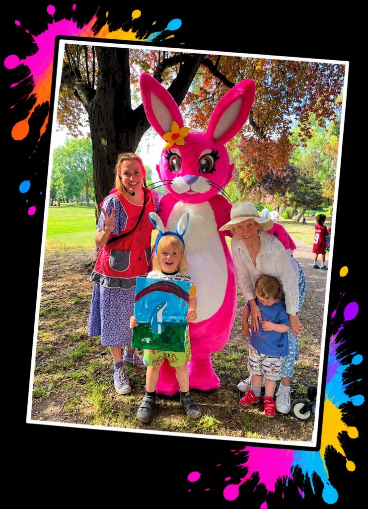 Families with the Pink Patsy mascot in the park, a little boy in bunny ears holding up his painting at a Painting in the Park with Pink Patsy event