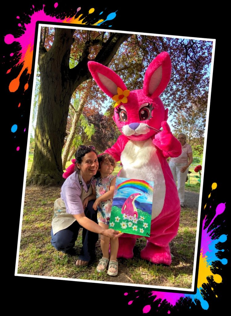 A mum and her little girl with the Pink Patsy mascot in the park, showing the rainbow painting they made at Painting in the Park with Pink Patsy