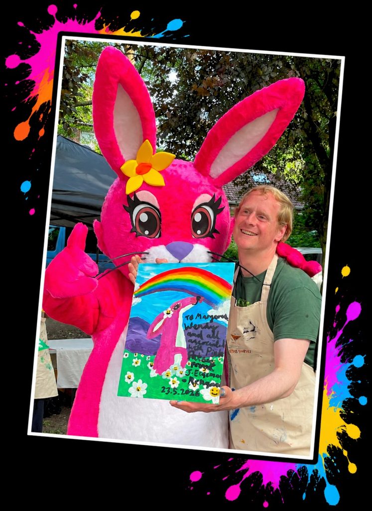 Jake and the Pink Patsy mascot giving a thumbs up with his Pink Patsy rainbow painting at a Painting in the Park event