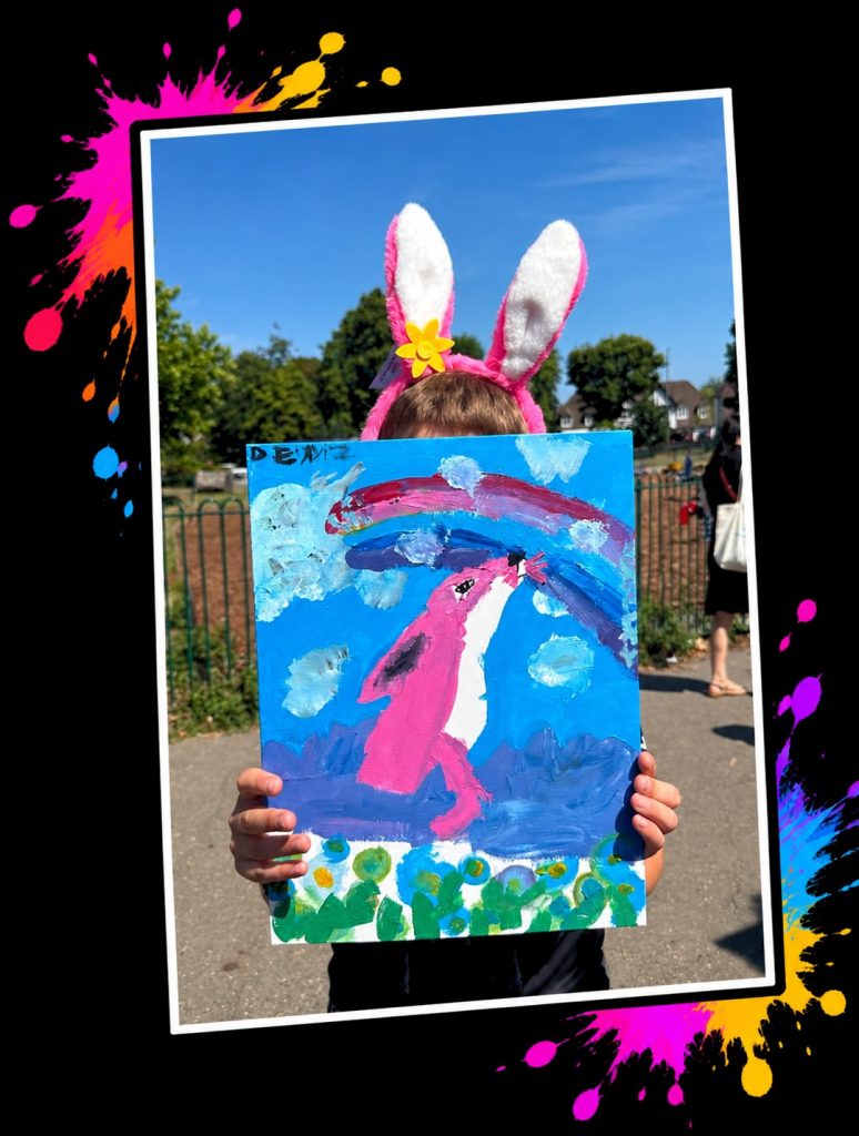 A young artist in Pink Patsy bunny ears holding up a painting of Pink Patsy under a rainbow at a Painting in the Park event