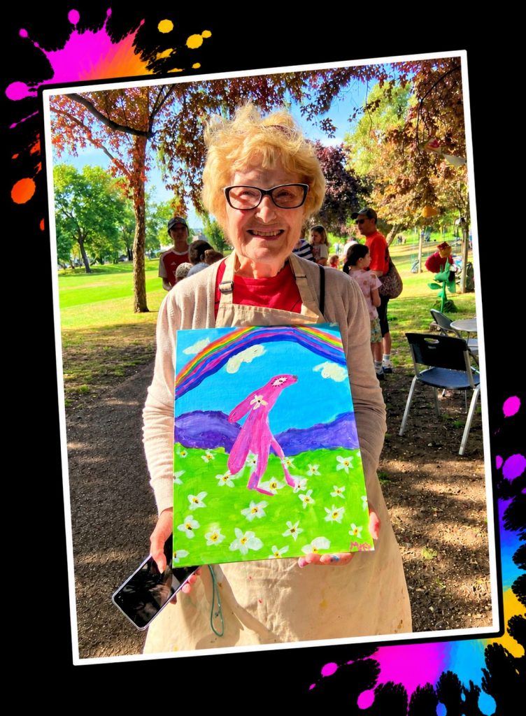 A smiling grandma in a painting apron holding her Pink Patsy rainbow painting at a Painting in the Park event, showing the sessions are for all ages