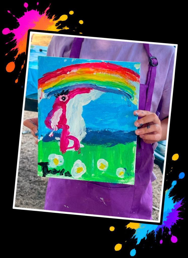 A young artist in a purple apron holding a bright painting of Pink Patsy under a big rainbow at a Painting in the Park event