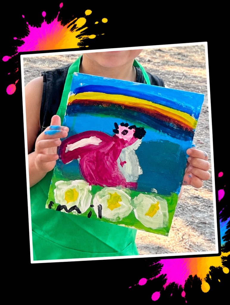 A young artist in a green apron holding a painting of Pink Patsy under a rainbow with big daisies at a Painting in the Park event