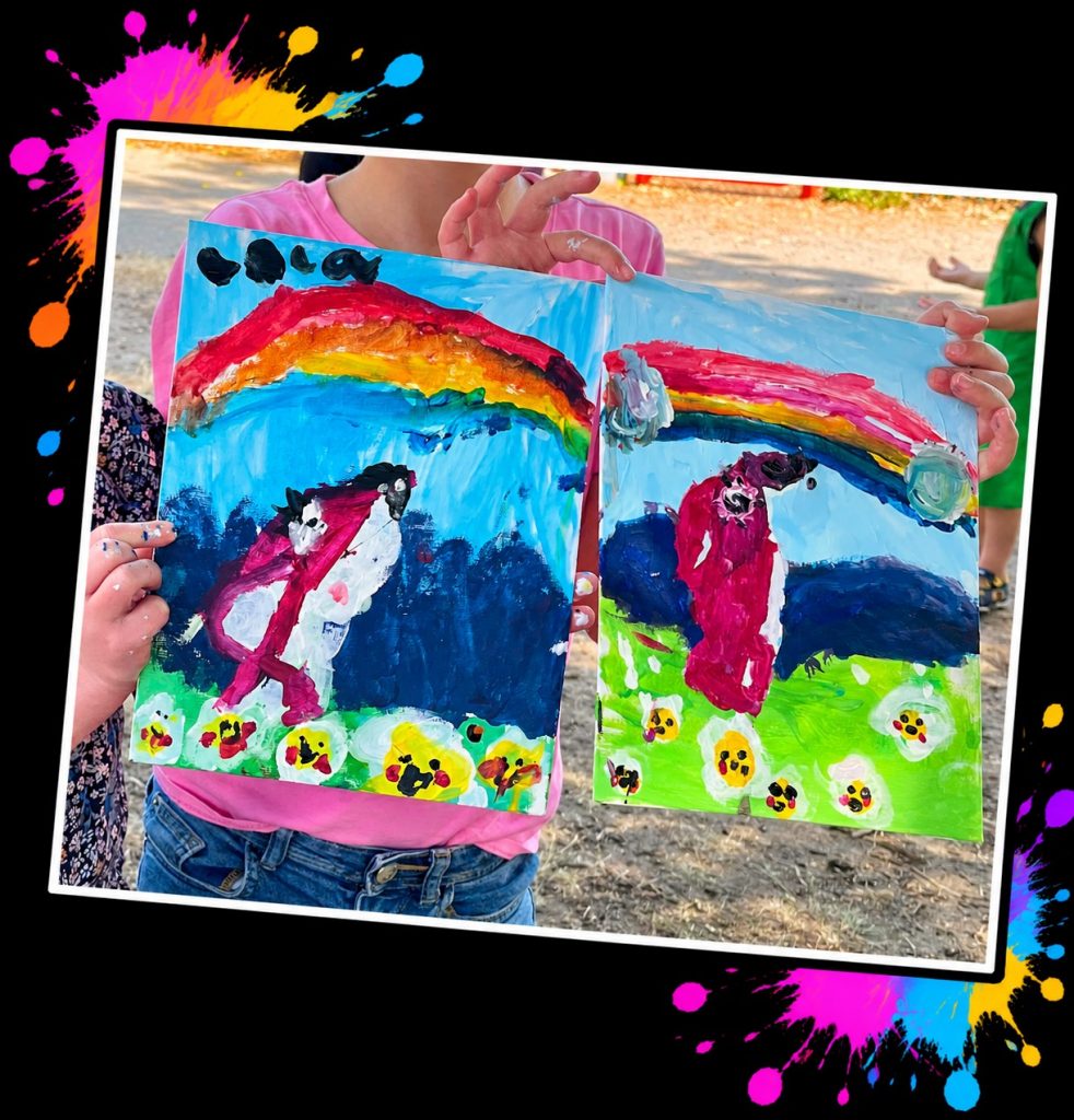 Two young artists holding up their colourful Pink Patsy rainbow paintings side by side at a Painting in the Park event