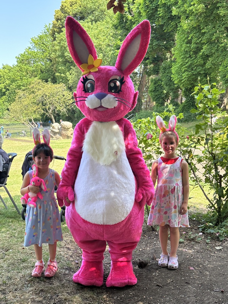 Two little girls in bunny ears and face paint with the Pink Patsy mascot at a Pink Patsy painting party in the park