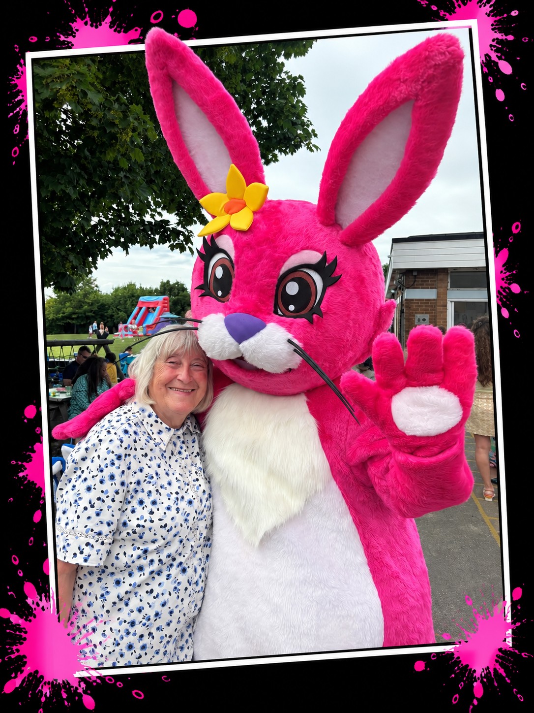 The Pink Patsy mascot, a giant pink teddy hare with a yellow flower, waving and hugging a visitor at a school event