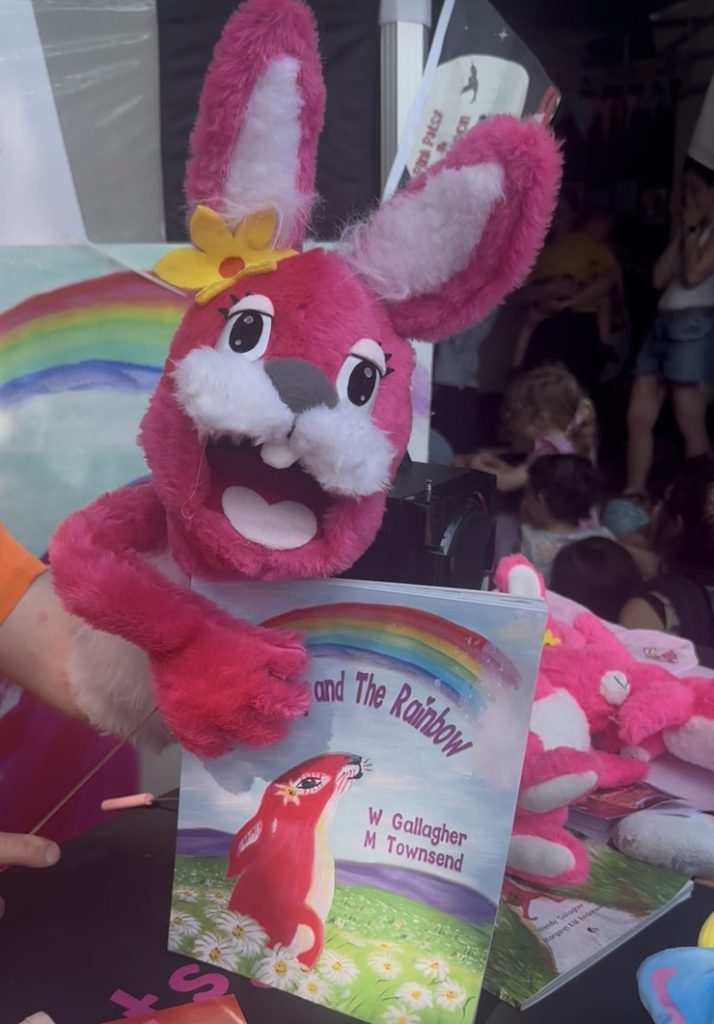 The Pink Patsy puppet holding the book Pink Patsy and the Rainbow at a live Pink Patsy Puppet Show