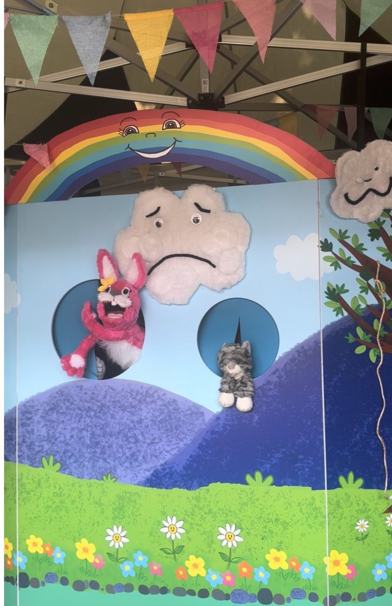 The Pink Patsy Puppet Show theatre at East Finchley Festival, with a smiling rainbow, sad cloud, Pink Patsy and the cat puppet under festival bunting