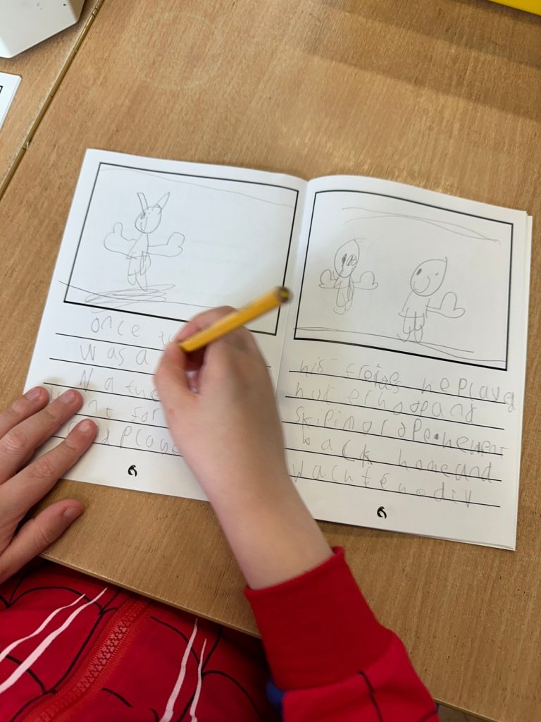 A child writing their own Pink Patsy story in the booklet, with pencil drawings of Pink Patsy and friends