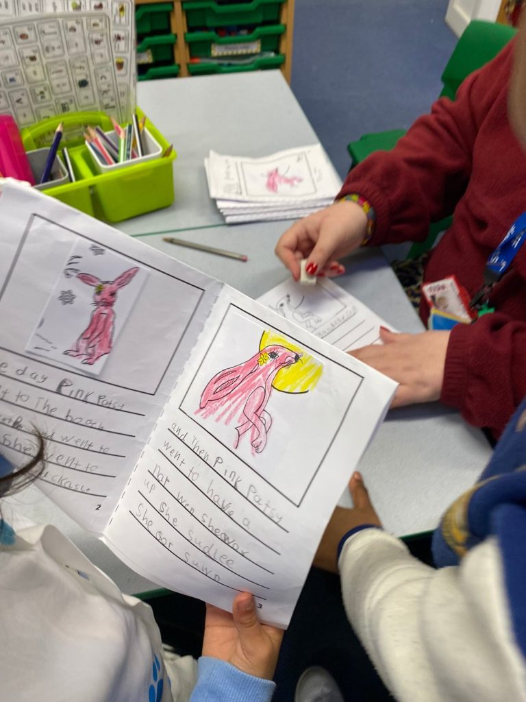 Children at a school table holding their own Pink Patsy story booklet, coloured pictures and handwritten story pages