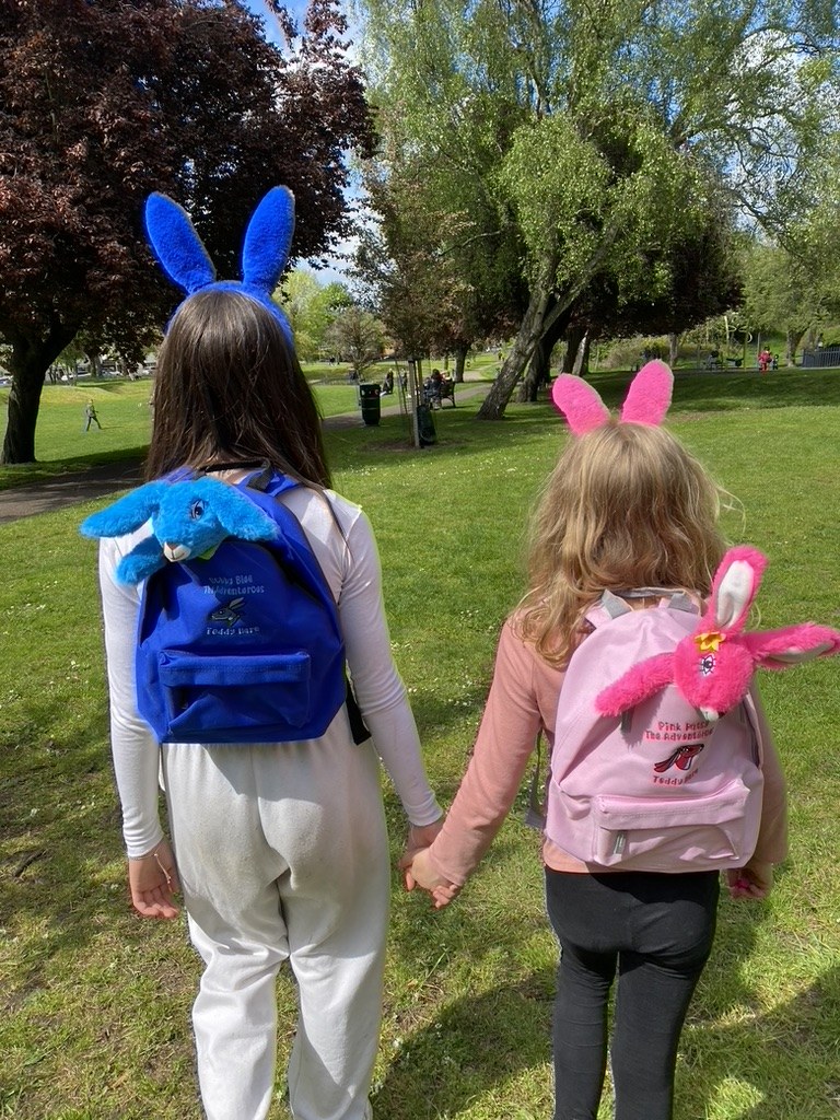 Two children walking in the park wearing the Pink Patsy and Bobby Blue adventure backpacks with the teddy hares