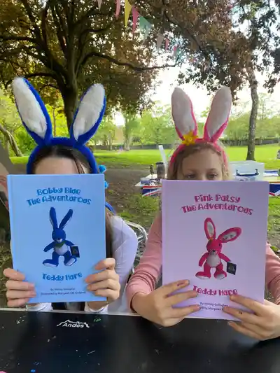 Two children in Bobby Blue and Pink Patsy ears holding up their Teddy Hare books in the park
