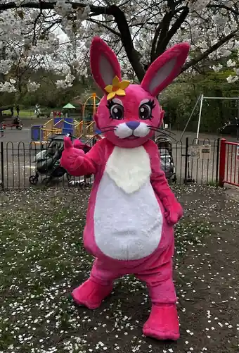 The Pink Patsy mascot, a giant pink hare with a yellow flower, giving a thumbs up in a park under cherry blossom
