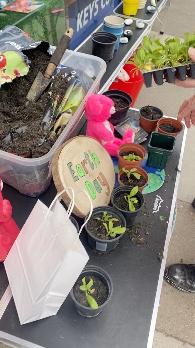 The Earth Day planting table with compost, a trowel, pots of seedlings, Pink Patsy and an Earth Day sign